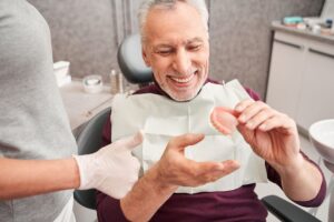 Man in dentists chair holding dentures smiling next to dentist giving thumbs up